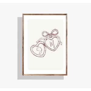May include: A framed print featuring a minimalist line drawing of two heart-shaped pendants connected by a bow. The pendants are outlined in burgundy, with the letters "M" and "D" inside. The background is off-white.