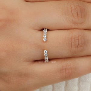 May include: An open-style gold ring featuring two small, vertical bars, each set with three clear, round-cut stones. The ring is positioned between the index and middle fingers, showcasing a minimalist design with a polished metal finish.