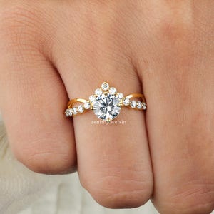 May include: A gold ring with a large, round, clear gemstone at the center. The band features smaller, clear gemstones and a decorative floral design. The ring is held up by a hand, showcasing the ring's intricate details.