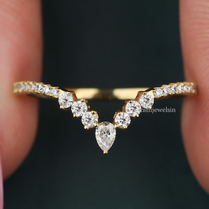 May include: A gold chevron-shaped ring featuring a series of small round-cut white stones along the band, culminating in a single pear-cut white stone at the center point. The band has a polished finish and a V-shaped design.