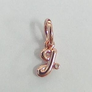Puede incluir: Un dije de oro rosa con la forma de la letra "J". El dije tiene un pequeño lazo en la parte superior para sujetarlo a una pulsera o collar. Un pequeño acento redondo está en la parte inferior de la letra.