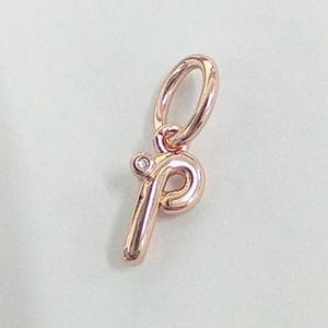 Könnte beinhalten: Roségoldener Anhänger in Form des Buchstabens "p". Der Anhänger hat oben eine kleine Schlaufe zum Befestigen an einem Armband oder einer Halskette. Oben auf dem Buchstaben ist ein kleiner, klarer Edelstein gesetzt.
