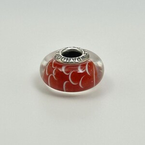 May include: A red and white glass bead with a silver-coloured metal core. The bead features a pattern of white curved shapes on a red background. The metal core is engraved with the word "PANDORA".