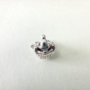 Puede incluir: Un charm Pandora plateado con forma redondeada y un peque&ntilde;o dise&ntilde;o en relieve en la parte superior. El charm tiene el logotipo de Pandora y otro texto grabado en su superficie. El charm es probablemente de plata de ley.