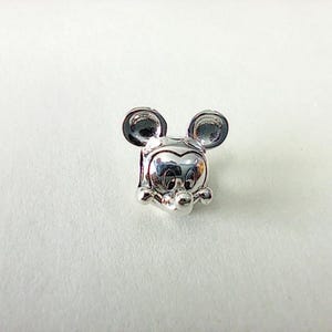 K&ouml;nnte beinhalten: Ein silberner Charm-Anh&auml;nger mit dem Gesicht von Mickey Mouse. Der Anh&auml;nger hat gro&szlig;e, runde Ohren, ein l&auml;chelndes Gesicht und kleine, runde H&auml;nde. Der Anh&auml;nger besteht aus gl&auml;nzendem, reflektierendem Metall.