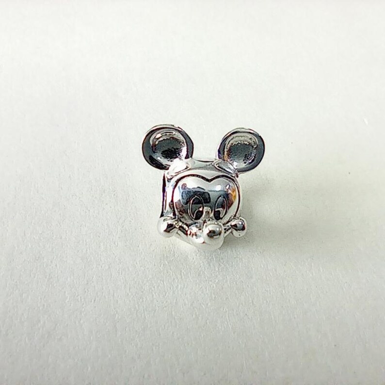 K&ouml;nnte beinhalten: Ein silberner Charm-Anh&auml;nger mit dem Gesicht von Mickey Mouse. Der Anh&auml;nger hat gro&szlig;e, runde Ohren, ein l&auml;chelndes Gesicht und kleine, runde H&auml;nde. Der Anh&auml;nger besteht aus gl&auml;nzendem, reflektierendem Metall.