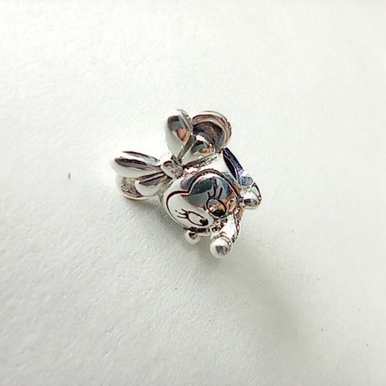 Puede incluir: Un charm de plata con forma de personaje de dibujos animados con un lazo en la cabeza. El charm tiene una cara detallada con ojos grandes y una nariz peque&ntilde;a. El charm es de metal brillante, probablemente una joya.