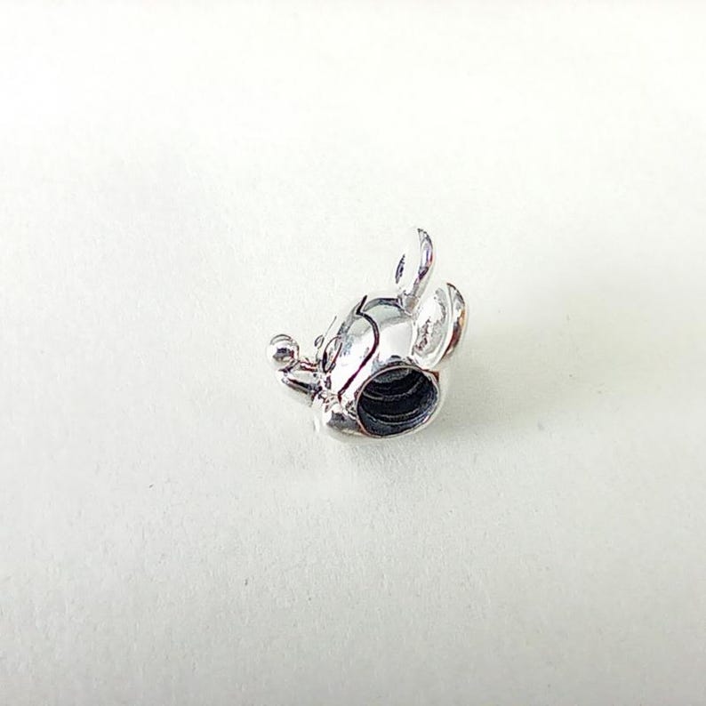 K&ouml;nnte beinhalten: Ein silberfarbener Charm in Form eines M&auml;usekopfes mit abgerundeten Ohren, einer kleinen Nase und einer kreisf&ouml;rmigen &Ouml;ffnung. Der Charm ist sehr detailliert und scheint aus Metall zu sein. Das Design erinnert an eine beliebte Zeichentrickfigur.