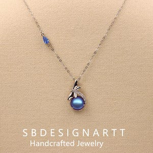 May include: A silver necklace with a dragonfly pendant and a blue pearl. The pendant is accented with small clear stones. A small blue teardrop-shaped stone is set on the chain. The text "SB DESIGNARTT Handcrafted Jewelry" is at the bottom.