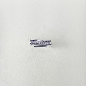 May include: A small, rectangular silver charm with a row of six clear, round gemstones. The charm has a slightly curved shape and is set against a plain white background. The gemstones are evenly spaced.