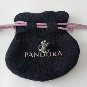 Puede incluir: Un charm plateado con forma de personaje de dibujos animados se muestra sobre una bolsa negra. La bolsa tiene la palabra "PANDORA" impresa en blanco. Un cord&oacute;n rosa es visible en la parte superior de la bolsa.