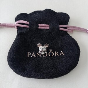 K&ouml;nnte beinhalten: Ein silberner Mickey Mouse-Charm liegt auf einem schwarzen Pandora-Beutel mit rosa Kordelzug. Der Beutel hat das Pandora-Logo in Silber aufgedruckt. Der Charm zeigt Mickeys Gesicht und Ohren.