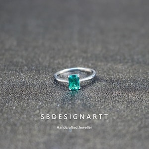 Peut inclure: Une bague en argent ornée d'une pierre précieuse rectangulaire vert émeraude. La bague est posée sur une surface gris foncé. Le texte "SBDESIGNARTT" et "Handcrafted Jeweller" sont visibles.