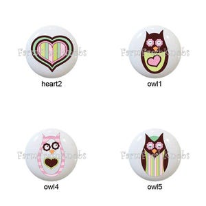 May include: A collection of eight white ceramic cabinet knobs. Designs include hearts and owls in various colors like brown, pink, green, and yellow. Some knobs have text that reads "heart1", "heart2", "owl1", "owl2", "owl3", "owl4", "owl5", and "wordart1".