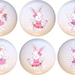 May include: Six white ceramic knobs with a pink and purple ballerina bunny rabbit design. Each knob features a different bunny rabbit pose.