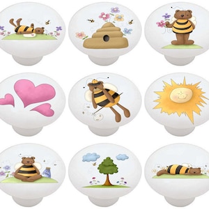 May include: Set of 9 ceramic cabinet knobs featuring a whimsical bee and bear theme. The knobs are white with colorful illustrations of bees, bears, hearts, a beehive, a sun, and a tree.