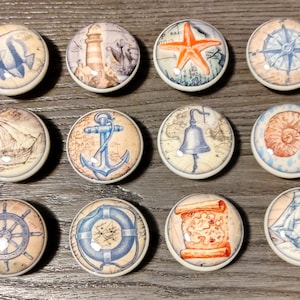 May include: Set of 10 round ceramic knobs with nautical themes. The knobs feature images of a lighthouse, a seashell, an anchor, a ship, a compass, a life preserver, a bell, a starfish, a seashell, and a ship's wheel.