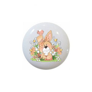 May include: A ceramic knob with a cartoon illustration of a brown bunny with a pink bow sitting in a field of flowers. A small pink bird is perched on the bunny's head.