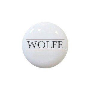 May include: A white, glossy, spherical object with the name "WOLFE" printed in black, centered horizontally. The text is framed by two thin, parallel brown lines. The background is plain white.