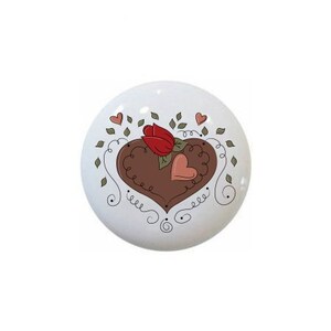 May include: A white ceramic knob with a brown heart design, two red flowers, and pink hearts. The heart is surrounded by black swirls and green leaves.