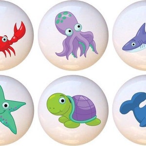 May include: Set of six ceramic knobs featuring cartoon sea creatures. The knobs are white with colorful cartoon images of a crab, octopus, shark, starfish, turtle, and whale.