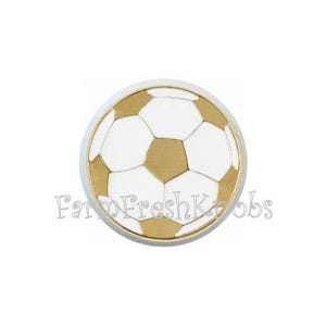 May include: A round cabinet knob with a soccer ball design. The knob features white and gold-colored pentagons and hexagons, with a gold-colored border. The text "Farm Fresh Knobs" is at the bottom.
