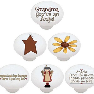 May include: Set of five white ceramic knobs with various designs. One knob features a brown star, another a yellow daisy, another a cartoon angel, another the text "Grandma you're an Angel", and the last one the text "Guardian Angels hear this prayer. Keep us in your loving care."