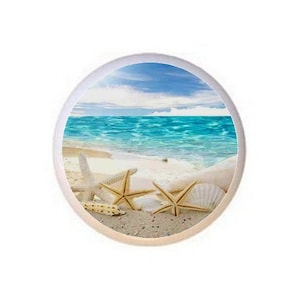 May include: A round white ceramic coaster with a beach scene featuring blue water, white sand, and three starfish. The starfish are arranged in a row on the sand.
