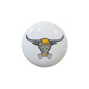 May include: A white ceramic button with a cartoon illustration of a gray bull's head with yellow horns and a bone in its mouth.