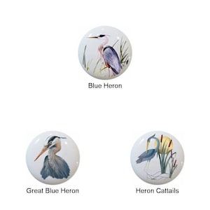 May include: Three white ceramic cabinet knobs with heron illustrations. One knob features a blue heron with the text "Blue Heron." Another shows a Great Blue Heron. The third knob depicts a heron with cattails, labeled "Heron Cattails."