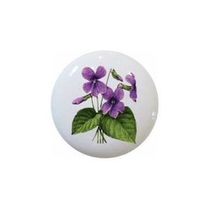 May include: A white, round ceramic cabinet knob featuring a botanical illustration of purple violets and green leaves. The floral design is centered on the knob, creating a decorative accent for furniture.