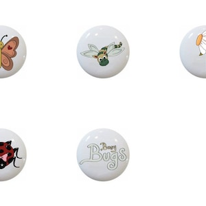 May include: A collection of ten white ceramic cabinet knobs. Each knob features a different insect or floral design, including a bee, butterfly, dragonfly, daisy, and ladybug. The text "Busy Bugs" is on one knob.