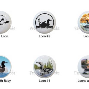 May include: A collection of six white ceramic cabinet knobs featuring loon bird designs. The knobs display various loon illustrations, including a flying loon, loons with babies, and loons at sunset. Each knob has the text "Flying Loon", "Loon #2", "Loon #6", "Loon with Baby", "Loon #1", and "Loons at Sunset".