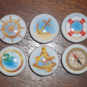 May include: Six nautical-themed ceramic cabinet knobs. Each knob features a different design: a ship's wheel, anchor, life preserver, porthole with island, sextant, and compass. The knobs are white with blue and gold accents.