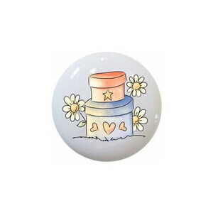 May include: A ceramic knob with a hand-painted design of two stacked boxes with hearts and a star on top, surrounded by three white daisies.