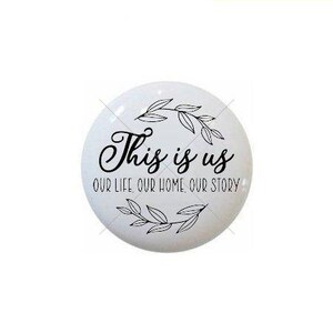 May include: A white, round decorative knob with black text and leaf accents. The text reads "This is us" above "Our life, our home, our story". The knob is likely for a cabinet or drawer.