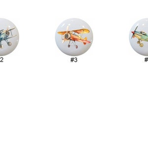May include: Six white ceramic knobs with different vintage airplane designs. Each knob features a different color scheme and airplane style.