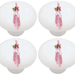 May include: Four white oval cabinet knobs. Each knob features a pair of pink ballet slippers hanging from a pink ribbon and a small floral arrangement. The knobs are a decorative accent for furniture.