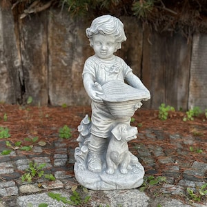 May include: A gray concrete garden statue of a young child holding a hat, with a dog at their feet. The statue is detailed with clothing and facial features. The statue is set on a stone base, with a wooden fence in the background.