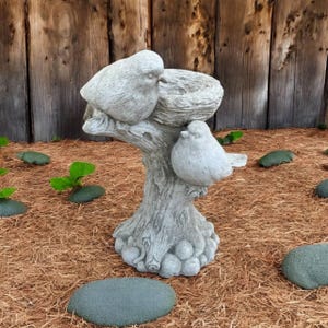 May include: A gray stone birdbath featuring a textured tree stump base with a nest on top and two sculpted birds perched on the sides. The surface of the sculpture has a weathered, mottled appearance with visible carved details on the birds and bark.