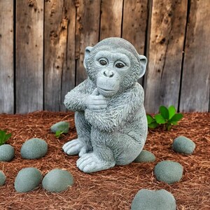 May include: A gray concrete monkey statue in a seated position, holding its hands together. The statue is surrounded by small, gray stones and sits on a bed of brown mulch. A wooden fence is in the background.