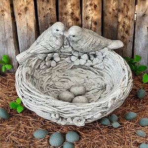 May include: A detailed, gray bird bath sculpture featuring two birds perched above a nest with three eggs. The nest is surrounded by flowers and sits on a bed of straw. Decorative stones are scattered around the base.