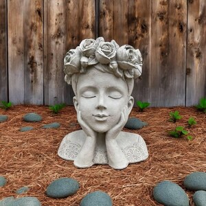May include: A serene, gray stone garden statue of a person with closed eyes, hands supporting the face, and a crown of roses. The statue is set in a garden bed with small rocks and greenery, against a wooden fence.