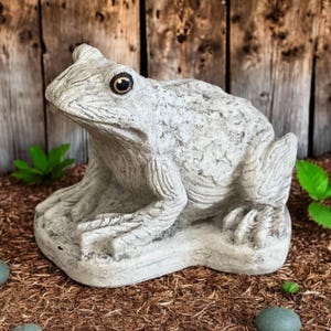 May include: A detailed, gray concrete frog statue with a textured surface. The frog has large, dark eyes and is sitting on a stone base. The statue is a garden ornament, suitable for outdoor decor.