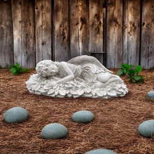 May include: A detailed, gray stone garden statue of a sleeping fairy with large wings, resting on a bed of leaves. The statue is set in a garden with a wooden fence background and several small, round, gray stones.