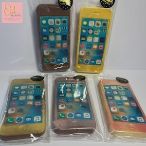 May include: Five individually wrapped soaps shaped like smartphones. The soaps are in shades of brown, yellow, gold, pink, and peach. Each soap features a detailed screen design with app icons and a date display. The soaps are packaged in clear plastic.
