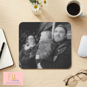 May include: A black and white family photo printed on a square mouse mat. The image shows a man holding a child, with a woman looking on. The mouse mat is on a light wood surface, with a coffee cup and flowers.