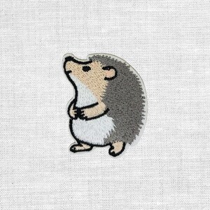 Könnte beinhalten: Gestickter Aufnäher mit einem Cartoon-Igel mit hellbraunem Gesicht und Bauch, grauem Rücken und schwarzen Augen. Der Igel steht aufrecht mit verschränkten Pfoten. Der Aufnäher hat einen weißen Rand und befindet sich auf weißem Stoffhintergrund.