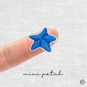 Könnte beinhalten: Ein kleiner, gestickter blauer Seestern-Aufnäher mit weißem Rand. Der Aufnäher wird von einem Finger gehalten, darunter steht in schwarzer Schrift "mini patch". Der Aufnäher ist ein DIY-Bastelartikel.