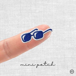 Könnte beinhalten: Ein kleiner, gestickter Aufnäher mit einer Sonnenbrille in Marineblau mit weißem Rand. Unter dem Aufnäher steht in Schreibschrift "mini patch".